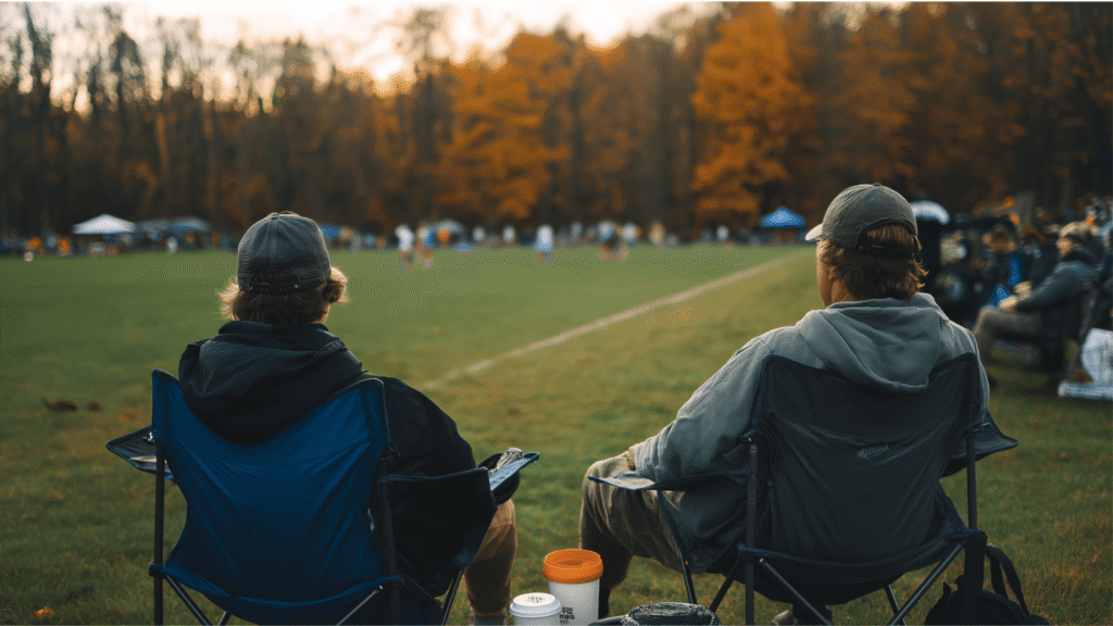 Two club lacrosse parents watching their kids play from folding chairs at a fall tournament